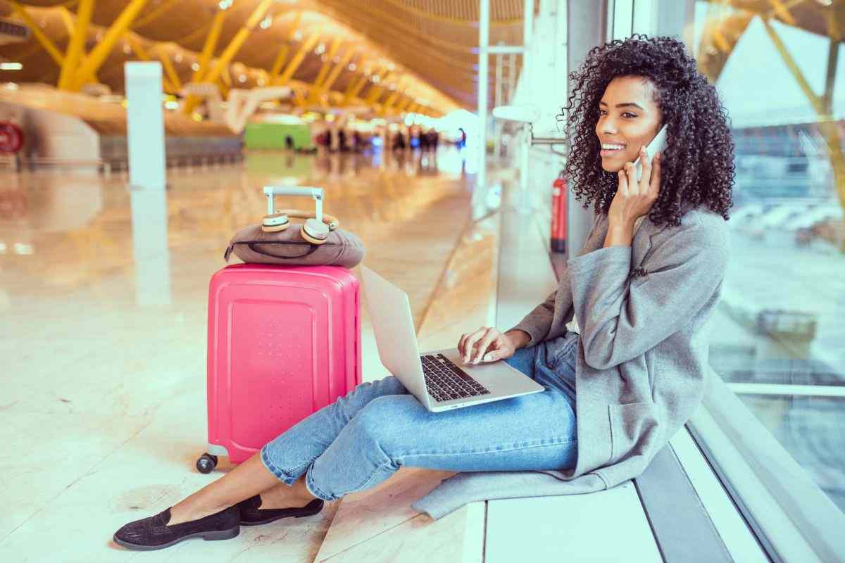 Donna seduta in un aeroporto con una valigia rosa accanto, che usa un laptop e parla al telefono
